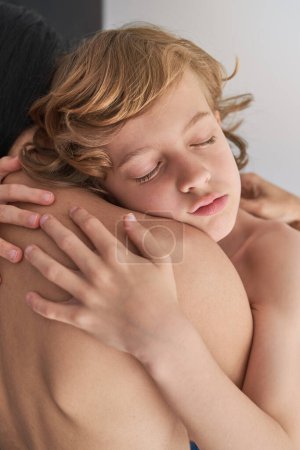 Photo for Peaceful kid curly hair and with closed eyes tenderly embracing anonymous topless mother while sitting in light room at home - Royalty Free Image