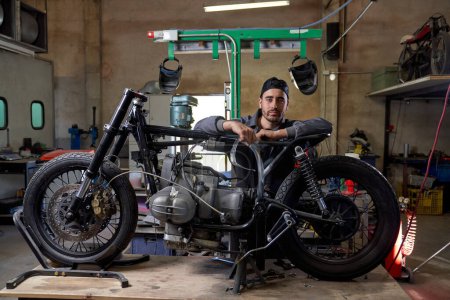 Foto de Grave mecánico barbudo en uniforme apoyado en una moto desmontada y mirando a la cámara en un taller de mala calidad - Imagen libre de derechos