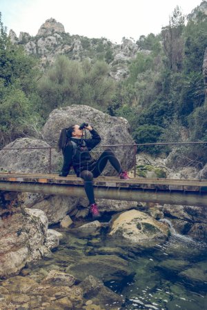 Foto de Cuerpo completo de joven viajera con pelo largo y oscuro en ropa casual sentada en pasarela y admirando la naturaleza a través de binoculares durante el trekking en valle montañoso de Mallorca - Imagen libre de derechos