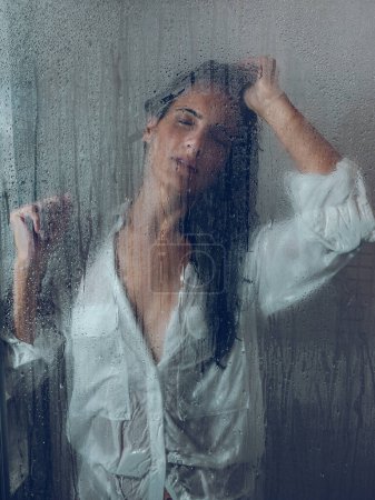 Téléchargez les photos : À travers un verre de jeune femme ethnique sensuelle avec de longs cheveux foncés en chemise blanche mouillée touchant la tête tout en se tenant dans la cabine de douche avec les yeux fermés - en image libre de droit