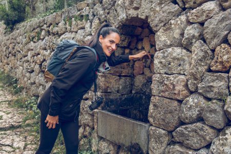 Foto de Vista lateral de la joven viajera hispana feliz con ropa de abrigo y mochila sonriendo y mirando a la cámara mientras enciende el agua de manantial que fluye del grifo en el nicho de piedra arqueada - Imagen libre de derechos