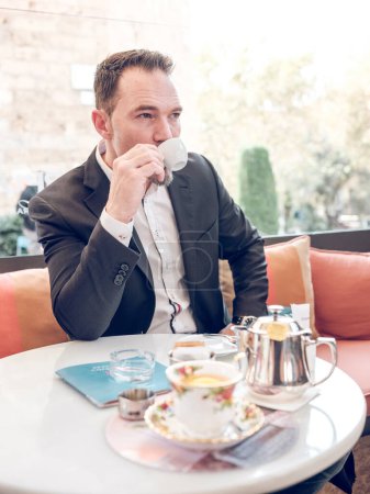 Photo for Self assured businessman in classy outfit sitting at table on summer terrace of cafe and enjoying espresso from cup while looking away - Royalty Free Image