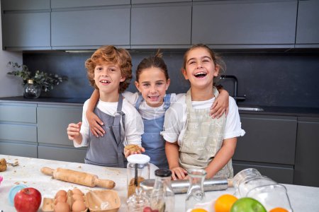 Téléchargez les photos : Enfants souriants dans des tabliers regardant la caméra à la table avec de la pâte à biscuits et divers ingrédients dans la cuisine légère à la maison - en image libre de droit