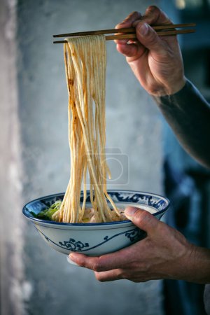 Téléchargez les photos : Recadrer un mâle tatoué anonyme avec des nouilles cuites sur des bâtonnets de nourriture sur un bol avec une délicieuse soupe japonaise - en image libre de droit