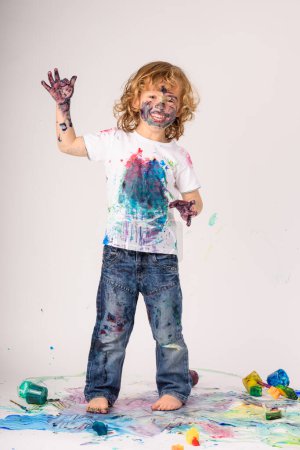 Photo for Full body of carefree barefoot dirty boy covered with colorful paints looking at camera while standing on messy floor with raised hand on white background - Royalty Free Image