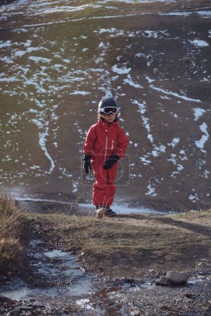 Téléchargez les photos : Enfant en vêtements de dessus et casque marchant sur un sol humide contre la pente enneigée de la montagne tout en passant une journée de printemps ensoleillée dans les hautes terres - en image libre de droit