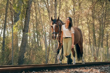 Photo for Content female equestrian leading chestnut horse along railway in forest on sunny day and looking away - Royalty Free Image