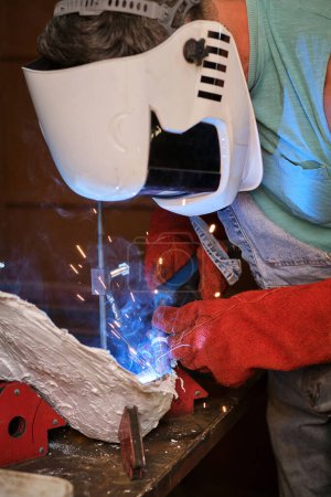 Foto de Artesano irreconocible en casco protector y guantes fijando detalle metálico con antorcha de soldadura mientras trabaja en taller profesional - Imagen libre de derechos