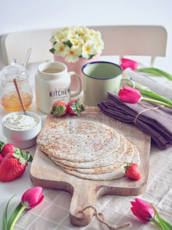 Photo for Tasty homemade crepes with chocolate pieces and strawberries surrounded decorative dishes placed on white table in kitchen - Royalty Free Image