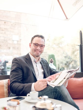 Téléchargez les photos : Homme adulte souriant en veste avec des lunettes de lecture journal tout en étant assis dans un café avec une tasse de café et téléphone et en regardant la caméra le matin - en image libre de droit