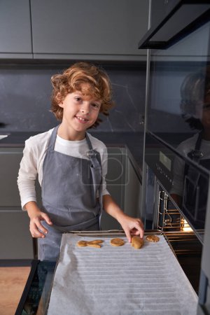 Photo for Smiling child in gray apron putting biscuits onto baking paper preparing to cook them in kitchen stove and looking at camera - Royalty Free Image