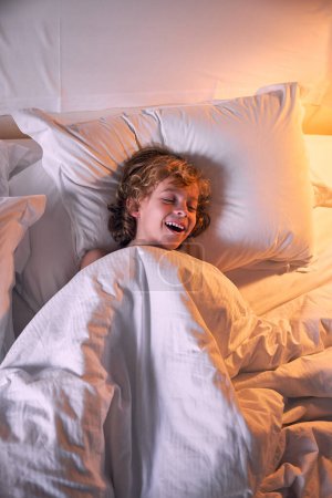 Téléchargez les photos : Du haut optimiste petit garçon couvert de couette chaude fermer les yeux et rire tout couché dans le lit la nuit à la maison - en image libre de droit
