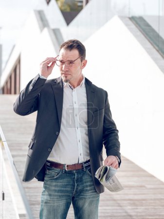 Téléchargez les photos : Entrepreneur masculin confiant dans des vêtements élégants avec journal détournant les yeux tout en mettant des lunettes tout en se tenant debout sur la rue ensoleillée près du bâtiment moderne - en image libre de droit