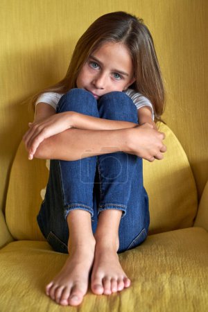 Photo for Full body of adorable barefoot child embracing knees and looking at camera while sitting in soft armchair in room - Royalty Free Image