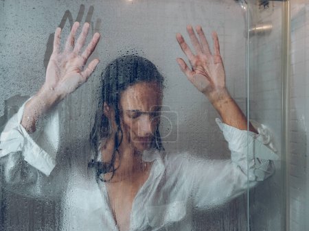 Foto de A través de cristal de triste hembra con pelo negro en ropa mojada tocando puerta transparente de cabina de ducha en baño ligero - Imagen libre de derechos