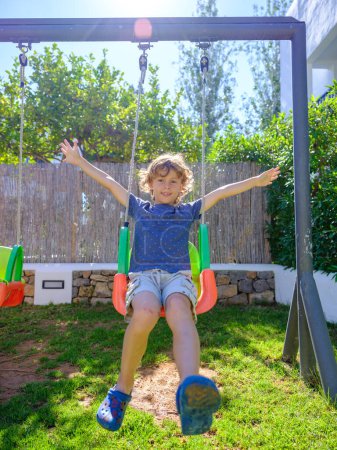 Photo for Full length of content young child with curly blond hair in casual clothes swinging on playground with outstretched arms and smiling while having fun in backyard during summer vacation - Royalty Free Image