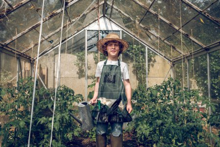 Photo for From below of positive preteen boy gardener in wicker hat and apron standing in hothouse among green plants with watering can - Royalty Free Image