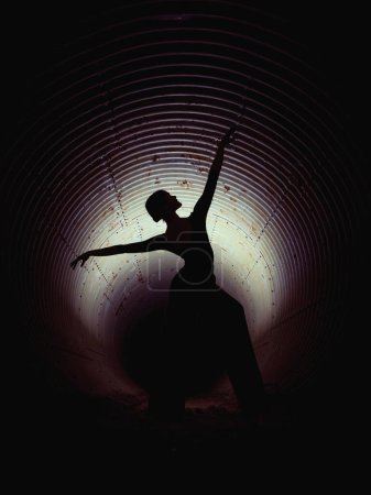 Téléchargez les photos : Silhouette de femme sans visage dansant les bras levés dans un tunnel souterrain sombre éclairé par la lumière - en image libre de droit