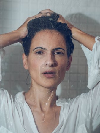 Téléchargez les photos : Brune vulnérable portant une chemise blanche recueillant les cheveux dans la cabine de douche tout en se mouillant et en regardant la caméra - en image libre de droit