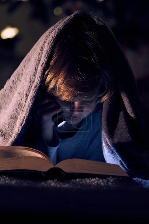 Téléchargez les photos : Focalisé garçon lecture livre intéressant avec lampe de poche tout en étant couché sur le lit sous plaid chaud dans la chambre noire la nuit - en image libre de droit