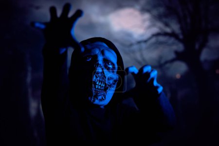 Téléchargez les photos : Monstre effrayant atteignant caméra avec griffe comme des mains tout en se faufilant à travers le cimetière sombre sous la lumière bleue néon la nuit - en image libre de droit