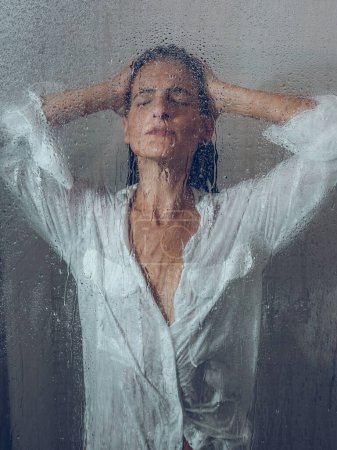 Photo for Through glass of charming female model with hand on head showering in transparent shower cabin with wet door during daily routine - Royalty Free Image