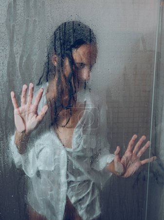 Photo for Through glass of sensitive female with wet hair touching transparent wet door while standing in shower cabin during daily routine - Royalty Free Image