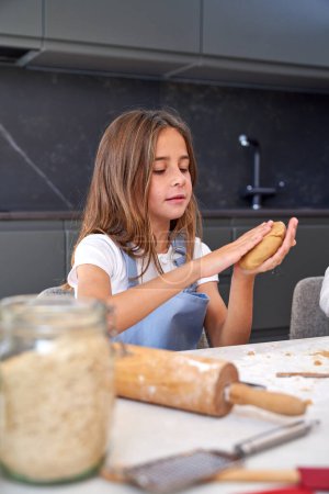 Photo for Attentive little girl with long hair in apron kneading soft dough while preparing delicious pie sitting at table in kitchen - Royalty Free Image