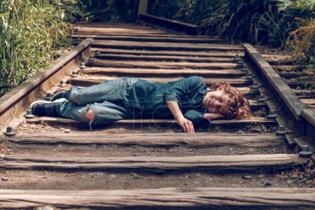 Photo for Full length of cheerful little kid with curly blond hair and closed eyes wearing denim clothes relaxing on wooden shabby railroad in sunny day - Royalty Free Image