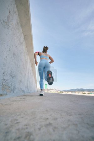 Foto de Desde abajo, vista trasera de una corredora femenina irreconocible corriendo durante el entrenamiento en la pasarela bajo el cielo azul - Imagen libre de derechos