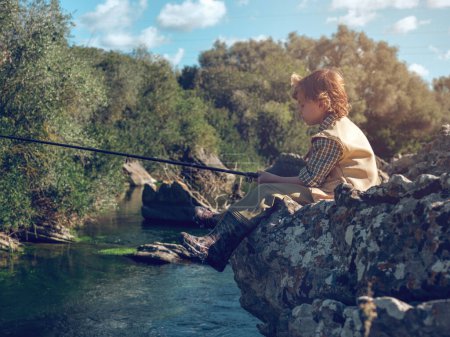 Téléchargez les photos : Vue latérale du garçon calme assis seul sur la rive du lac rocheux et la pêche avec canne sur fond de bois vert - en image libre de droit