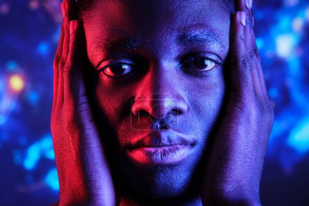 Foto de Cultivo triste afroamericano masculino con las manos en la cara contra luces de colores mirando a la cámara - Imagen libre de derechos