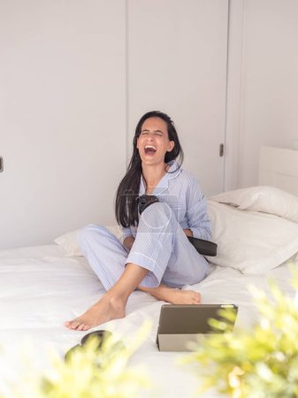 Foto de Cuerpo completo de hembra descalza en ropa de dormir sentada en la cama mientras ve fotos en la cámara profesional y se ríe - Imagen libre de derechos