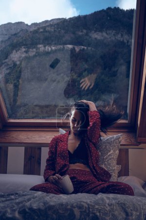 Foto de Mujer tranquila sentada en la cama y tocando el cabello mientras disfruta de fin de semana en acogedora casa de campo en las tierras altas - Imagen libre de derechos
