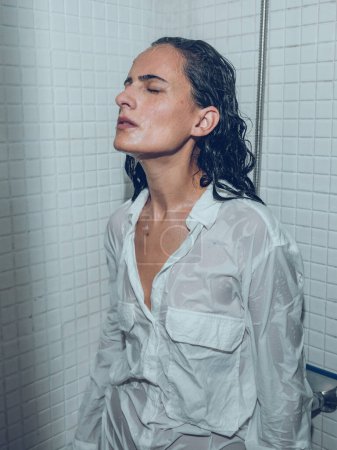 Foto de Hembra concentrada reflexiva en camisa blanca mojada parada debajo de agua que salpica en cabina de ducha y que tiene alivio de tensión - Imagen libre de derechos