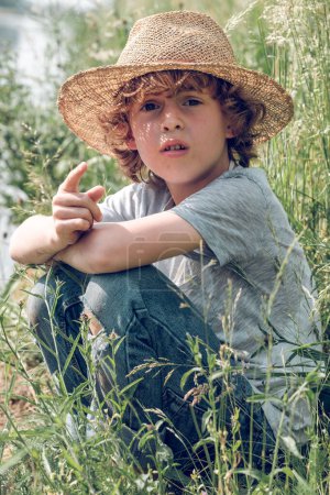 Photo for Charming kid with curly hair in straw hat sitting on green grass on channel coast and pointing finger while enjoying summer day and looking at camera - Royalty Free Image
