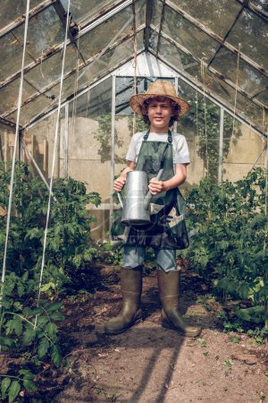 Téléchargez les photos : Corps complet de jardinier garçon préadolescent positif en bottes de gomme et chapeau en osier et tablier debout près des lits de jardin en serre - en image libre de droit