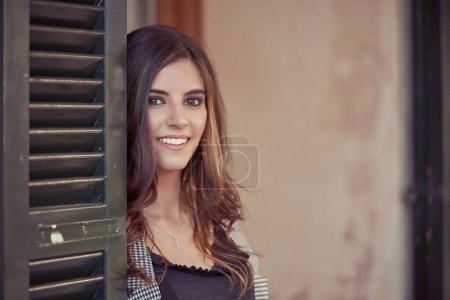 Foto de Encantadora hembra de cabello oscuro mirando a la cámara con sonrisa mientras está de pie cerca de la puerta negra del edificio en la calle de la ciudad - Imagen libre de derechos