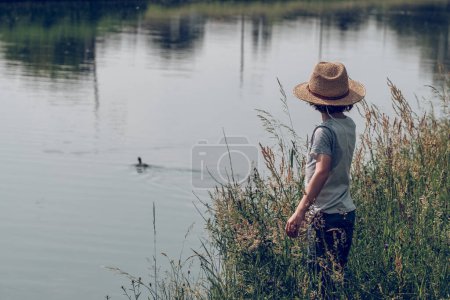 Photo for Side view of unrecognizable kid in straw hat standing among green grass on shore of channel and looking at duck on water in summer day - Royalty Free Image
