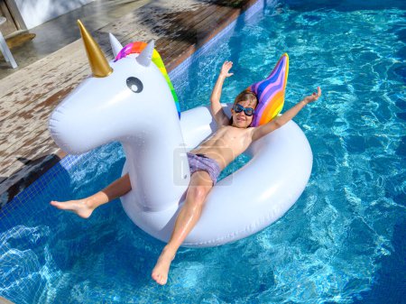 Photo for From above full body of shirtless excited little boy in shorts and diving goggles lying on unicorn shaped inflatable tube with raised arms and floating in outdoor pool in sunlight - Royalty Free Image
