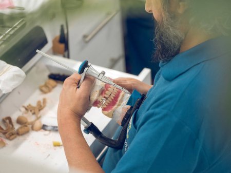 Téléchargez les photos : De dessus de vue latérale technicien principal de culture d'homme ajustant la prothèse dentaire de mâchoire dans l'articulateur tout en travaillant dans le laboratoire professionnel - en image libre de droit