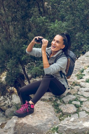Téléchargez les photos : Corps complet de voyageuse souriante avec sac à dos assis sur une falaise pierreuse et regardant des jumelles admirant la nature sur le terrain Biniaraix de la région de Soller à Majorque - en image libre de droit