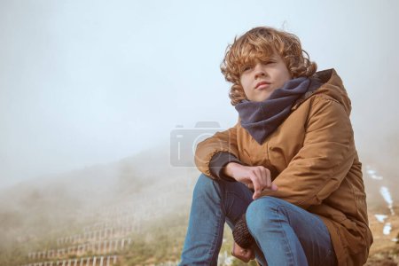 Téléchargez les photos : Serieux garçon aux cheveux bouclés en vêtements chauds assis sur le sol dans la vallée vallonnée couverte de vapeur et profitant du paysage - en image libre de droit