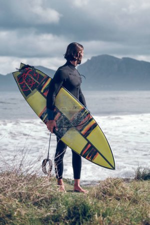 Photo for Full body of young muscular bearded male athlete in black wetsuit with surfboard in hand standing on grassy coast and looking away after practicing surfing in ocean against overcast sky - Royalty Free Image