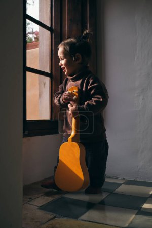 Téléchargez les photos : Curieux enfant avec instrument de musique regardant loin tout en se tenant sur le sol carrelé contre la fenêtre à la maison dans la lumière du soleil - en image libre de droit