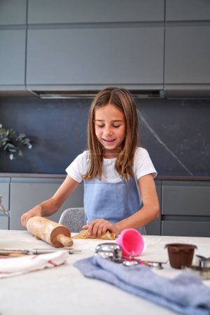 Photo for Optimistic girl in apron with wooden rolling pin kneading cookie dough while preparing for baking in light modern kitchen at home - Royalty Free Image
