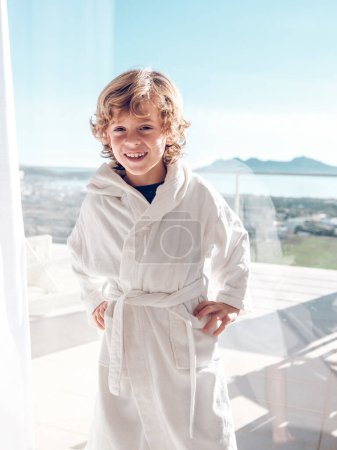 Téléchargez les photos : Par la fenêtre heureux garçon préadolescent en peignoir doux debout sur le balcon de la chambre de suite tout en se reposant à l'hôtel en vacances d'été à Pollensa ville de Majorque île - en image libre de droit