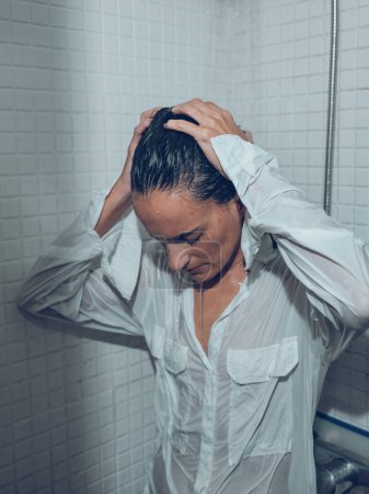 Photo for Fatigue female in wet shirt standing under splashing water in shower cabin and touching head - Royalty Free Image