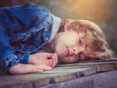 Téléchargez les photos : Enfant blond veste en denim avec les cheveux bouclés regardant la caméra tout en étant couché sur un chemin en bois dans la campagne - en image libre de droit