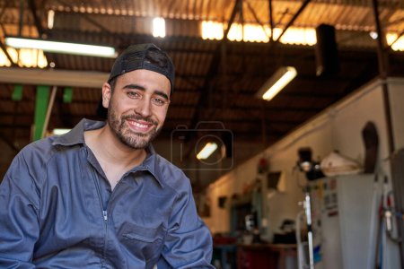 Foto de Mecánico masculino profesional en ropa de trabajo y gorra sonriendo y mirando a la cámara mientras está sentado en el garaje sobre fondo borroso - Imagen libre de derechos